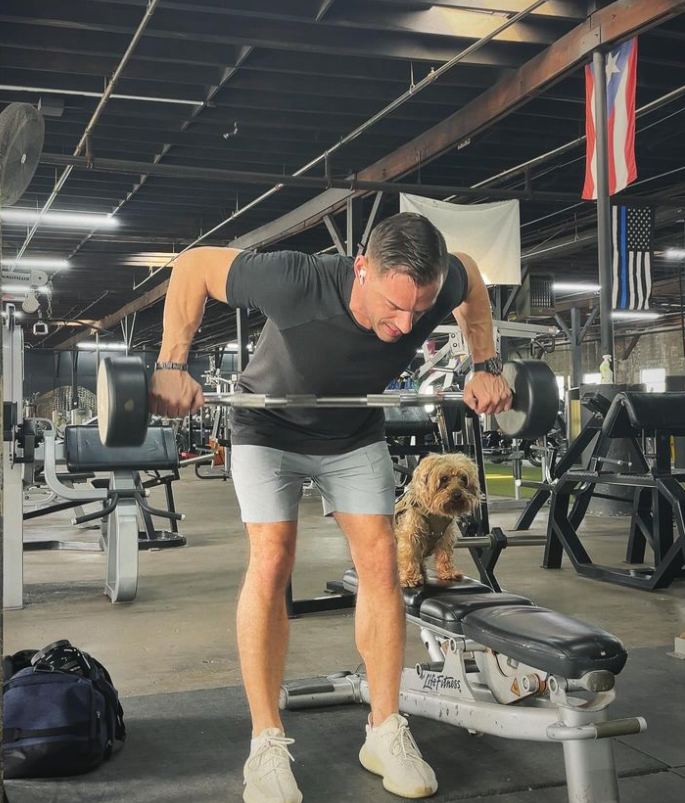 Man performing a dumbbell workout in a gym with a small dog sitting on a bench, representing a fitness routine for traveling professionals.