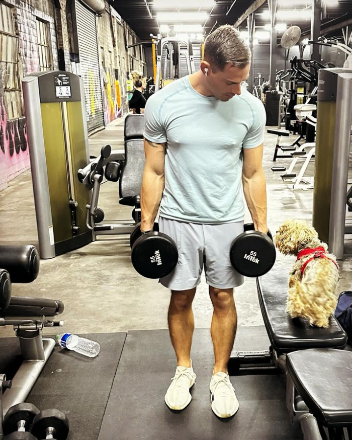 Man lifting dumbbells in a gym with a small dog on the bench, representing workout routines suitable for travel nurses, flight attendants, and pilots to maintain fitness while traveling.