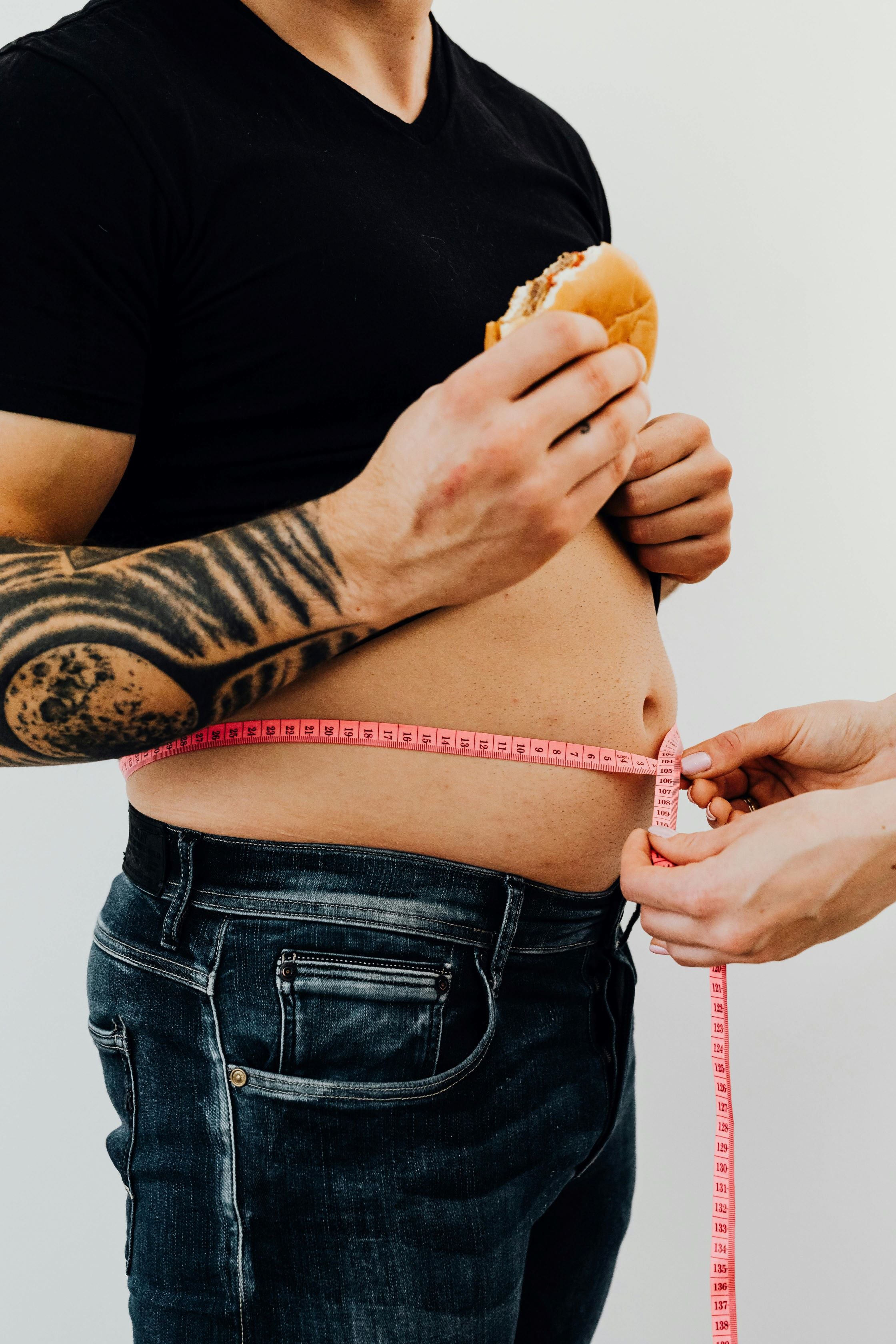 A close-up image of a person eating a burger while having their waist measured with a pink tape measure, symbolizing the start of a weight loss or fitness journey.